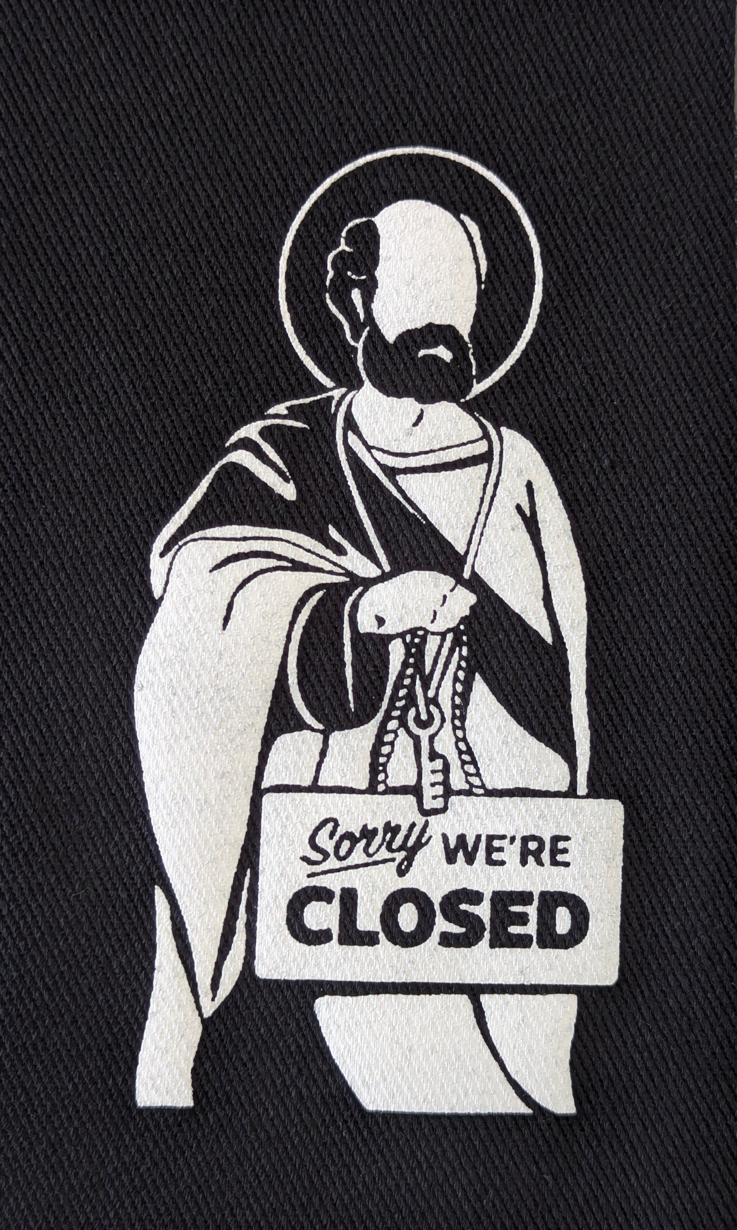 fabric patch of a Saint Peter holding a 'Sorry We're Closed' sign on black fabric.