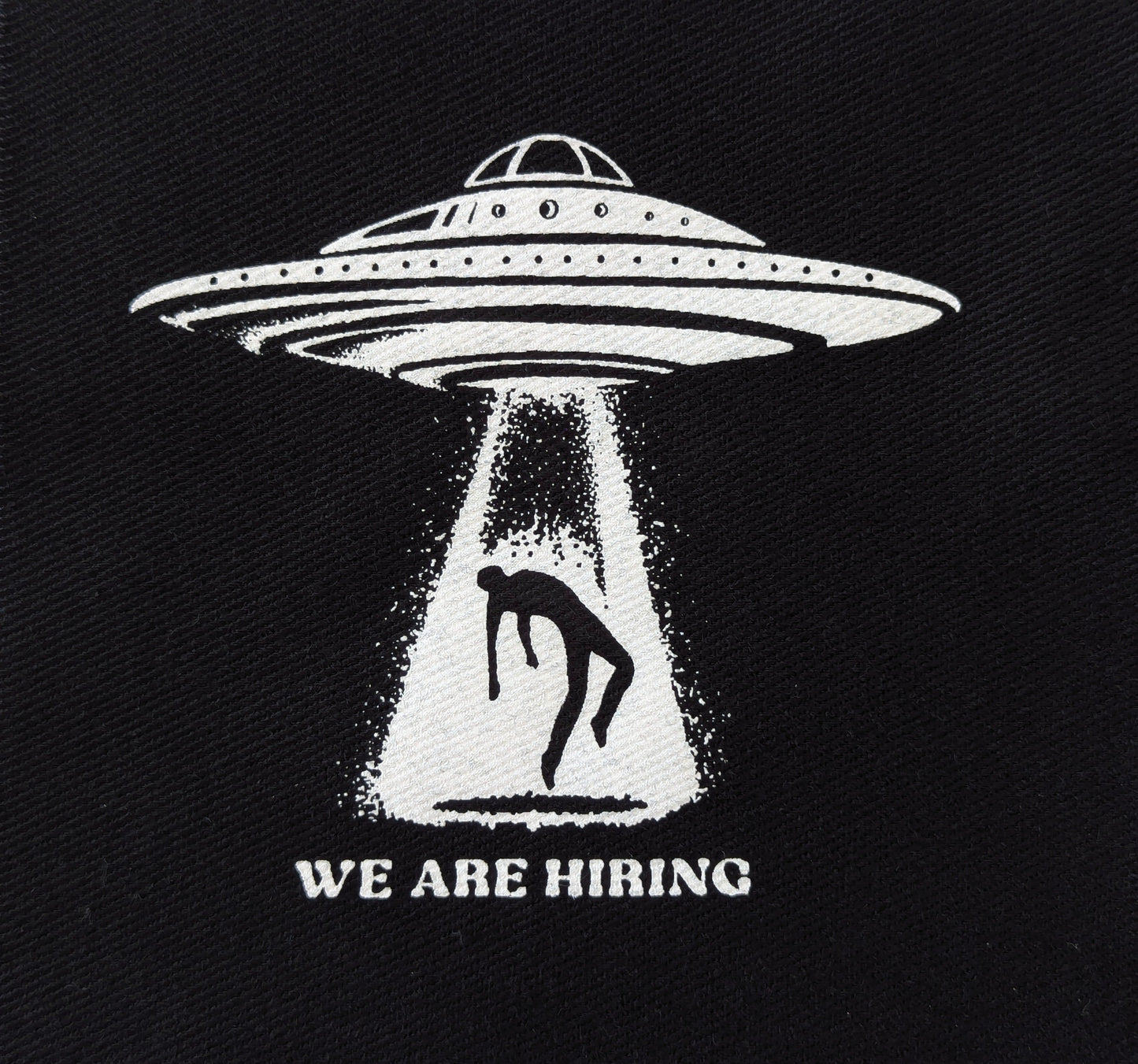 Black patch with white UFO and silhouette of a person, labeled 'WE ARE HIRING', on a dark background.