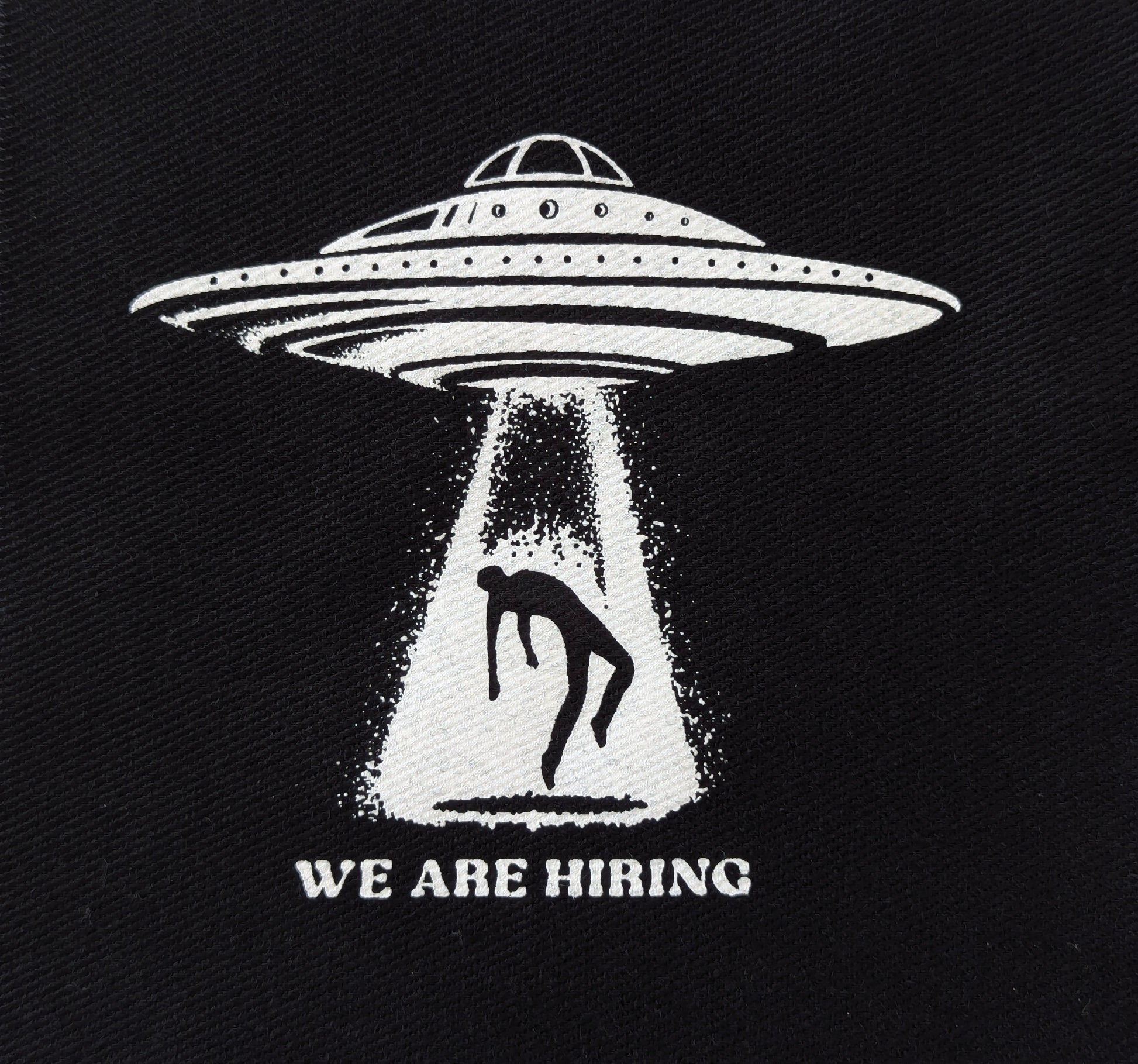 Black patch with white UFO and silhouette of a person, labeled 'WE ARE HIRING', on a dark background.